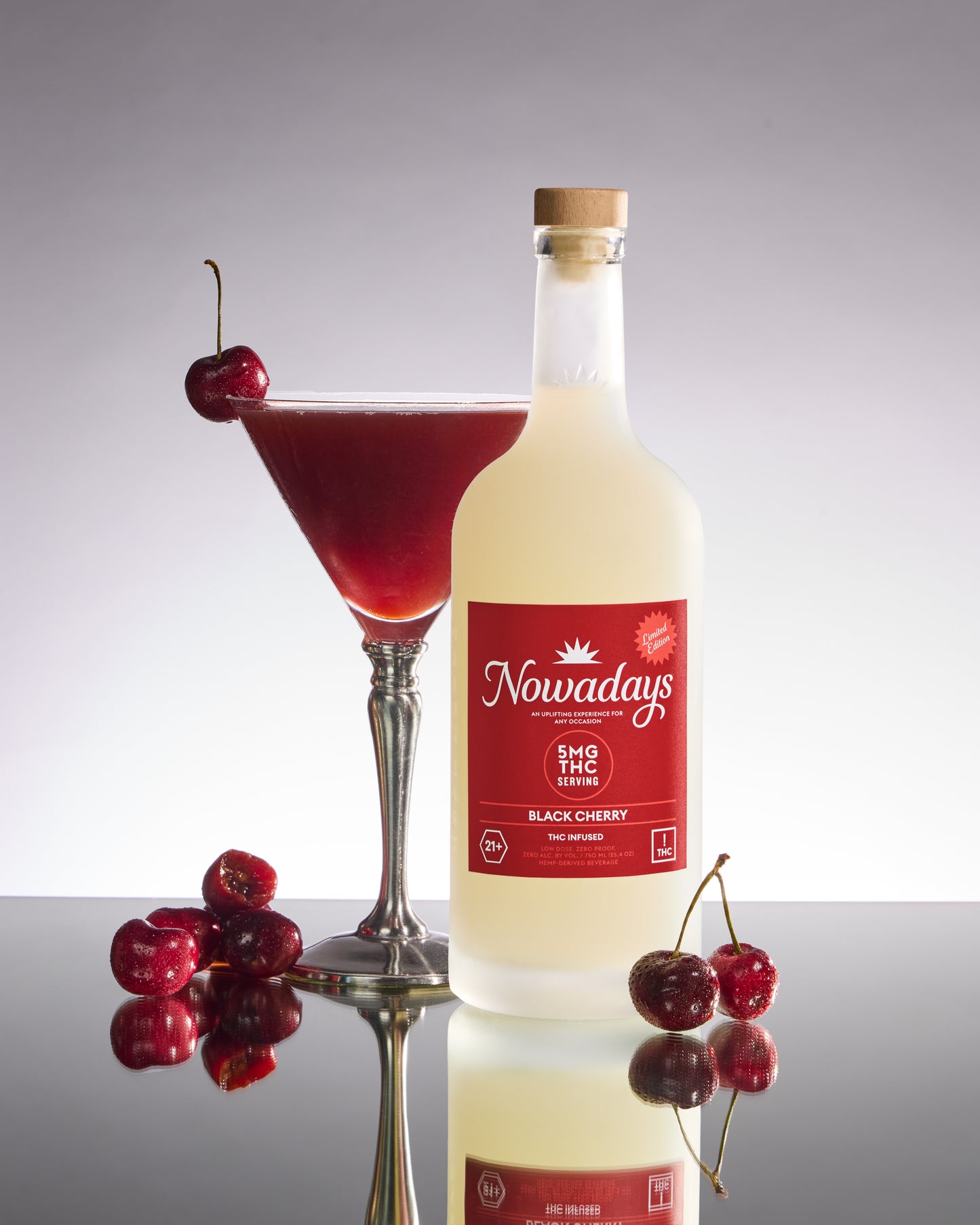 Bottle of Nowadays Black Cherry with a cocktail and cherries on a reflective surface.