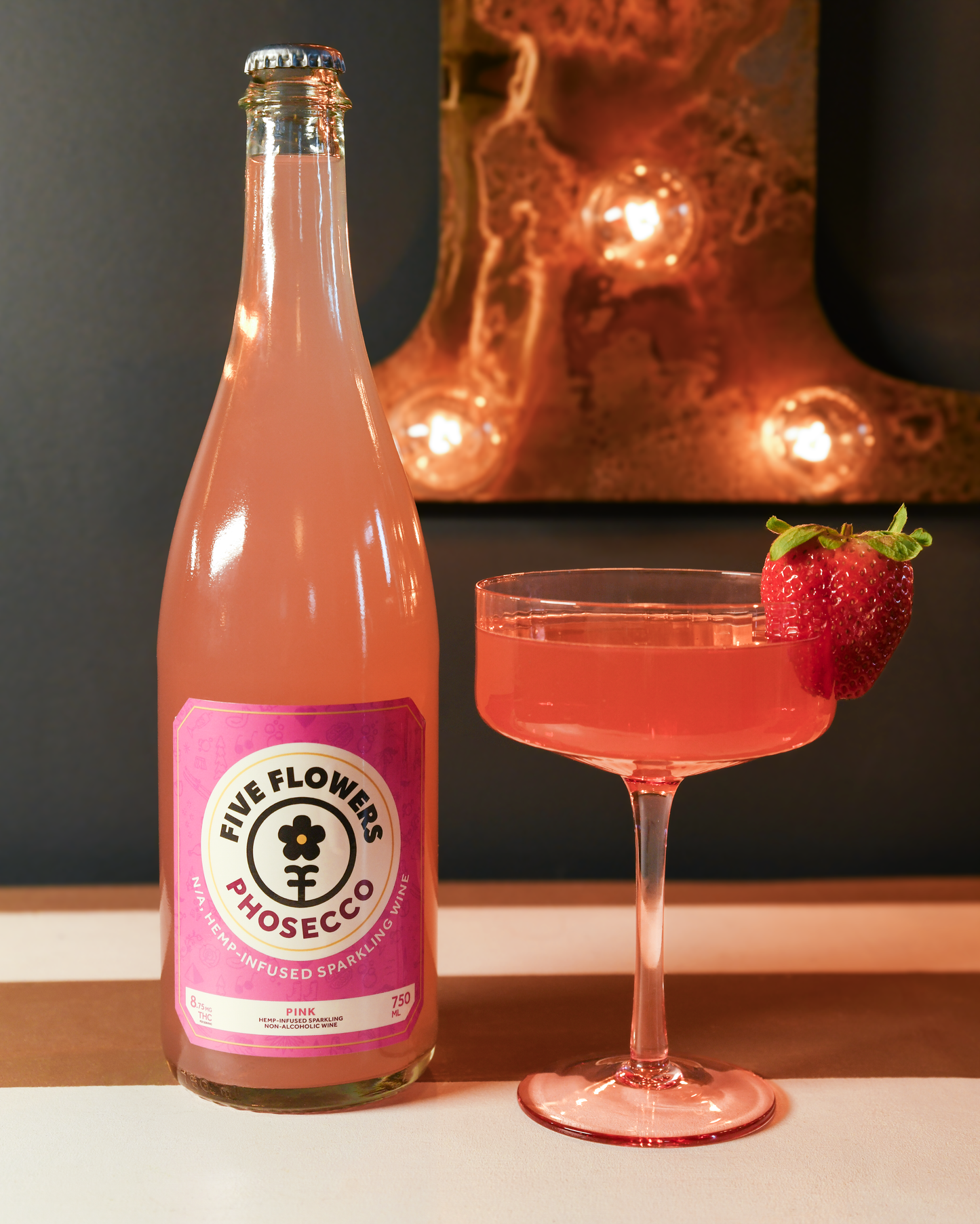 Bottle of Five Flowers Prosecco with a cocktail glass filled with pink liquid and a strawberry garnish, set against a dark background.