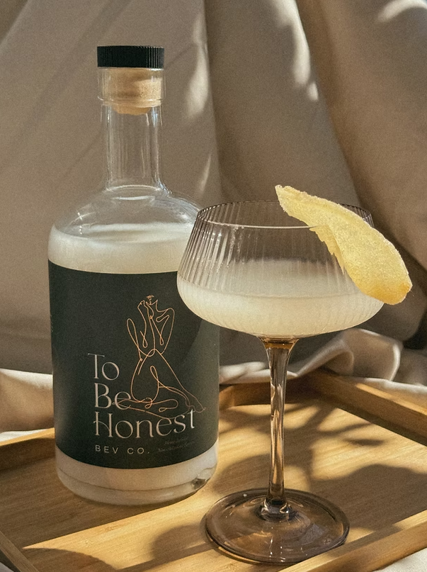 A bottle of 'To Be Honest' hemp infused non-alcoholic spirit next to a filled glass garnished with a slice of citrus, both placed on a wooden surface.