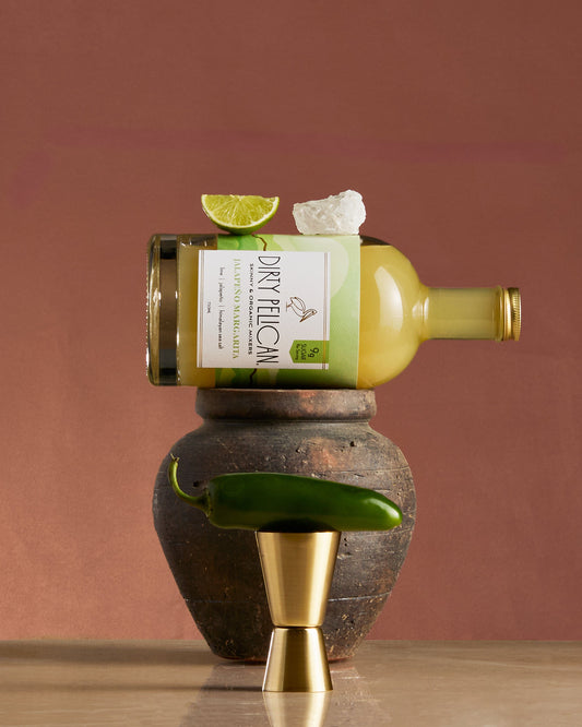 Bottle of 'Dirty Feliz' tequila with a lime wedge and jalapeño on a wooden and stone surface.