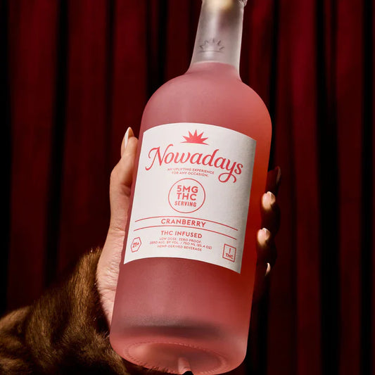 Pink bottle of 'Nowadays' cranberry THC-infused drink held by a hand against a red curtain background.